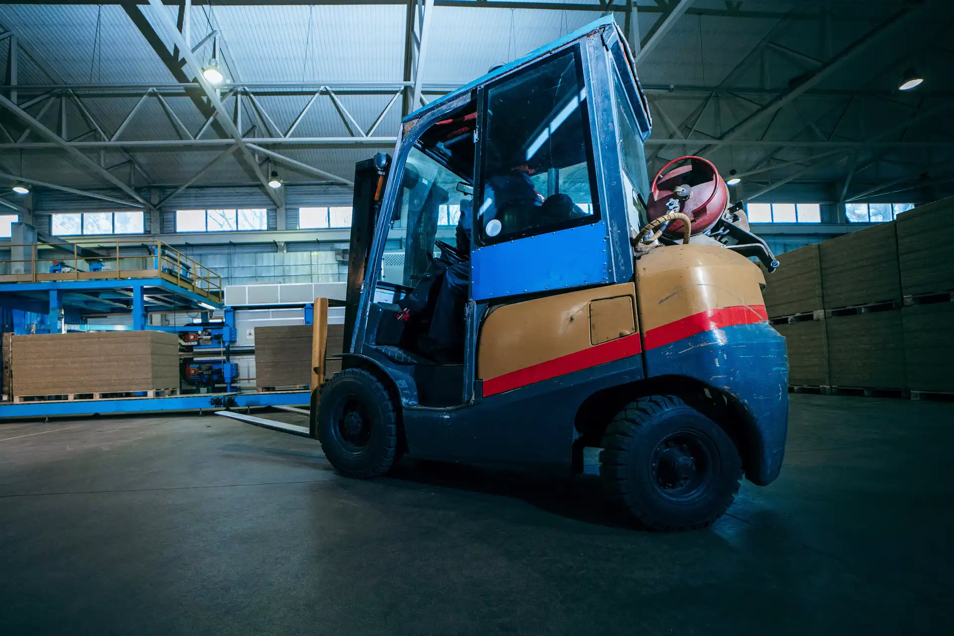 A yellow and blue forklift with a red propane tank sits in a dimly lit industrial warehouse with stacked materials.