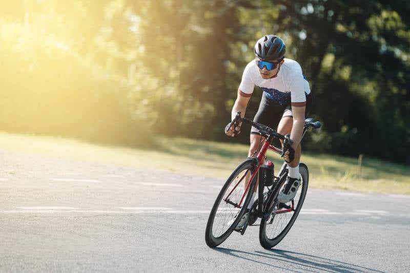 A cyclist in a helmet, sunglasses, and athletic wear rides a red road bike on a sunny road.