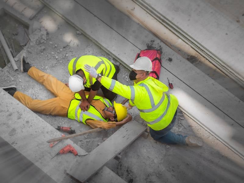Construction worker performing CPR on another worker lying on the ground at a construction site.