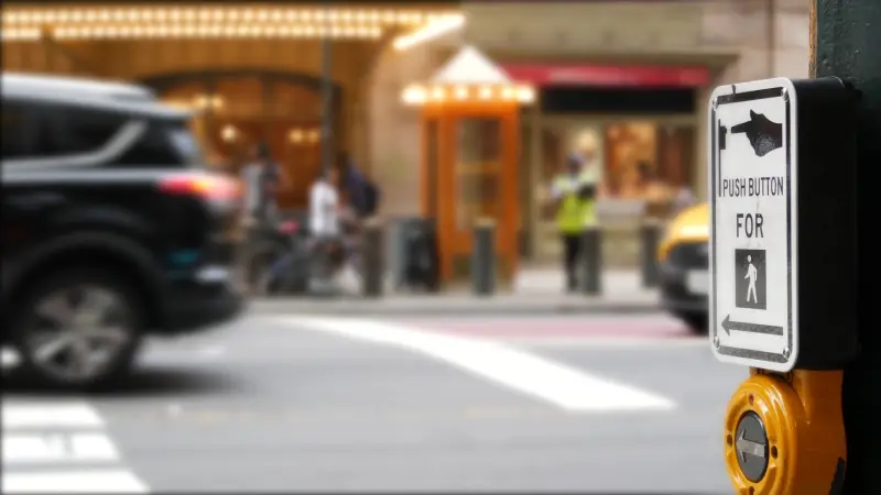 Pedestrian crosswalk button sign reading PUSH BUTTON FOR with a pedestrian icon, on a blurry city street.