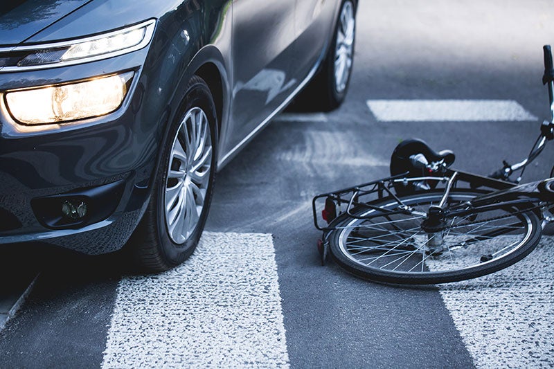 Dark gray car with headlights on, parked next to a fallen bicycle on a crosswalk, implying a collision.