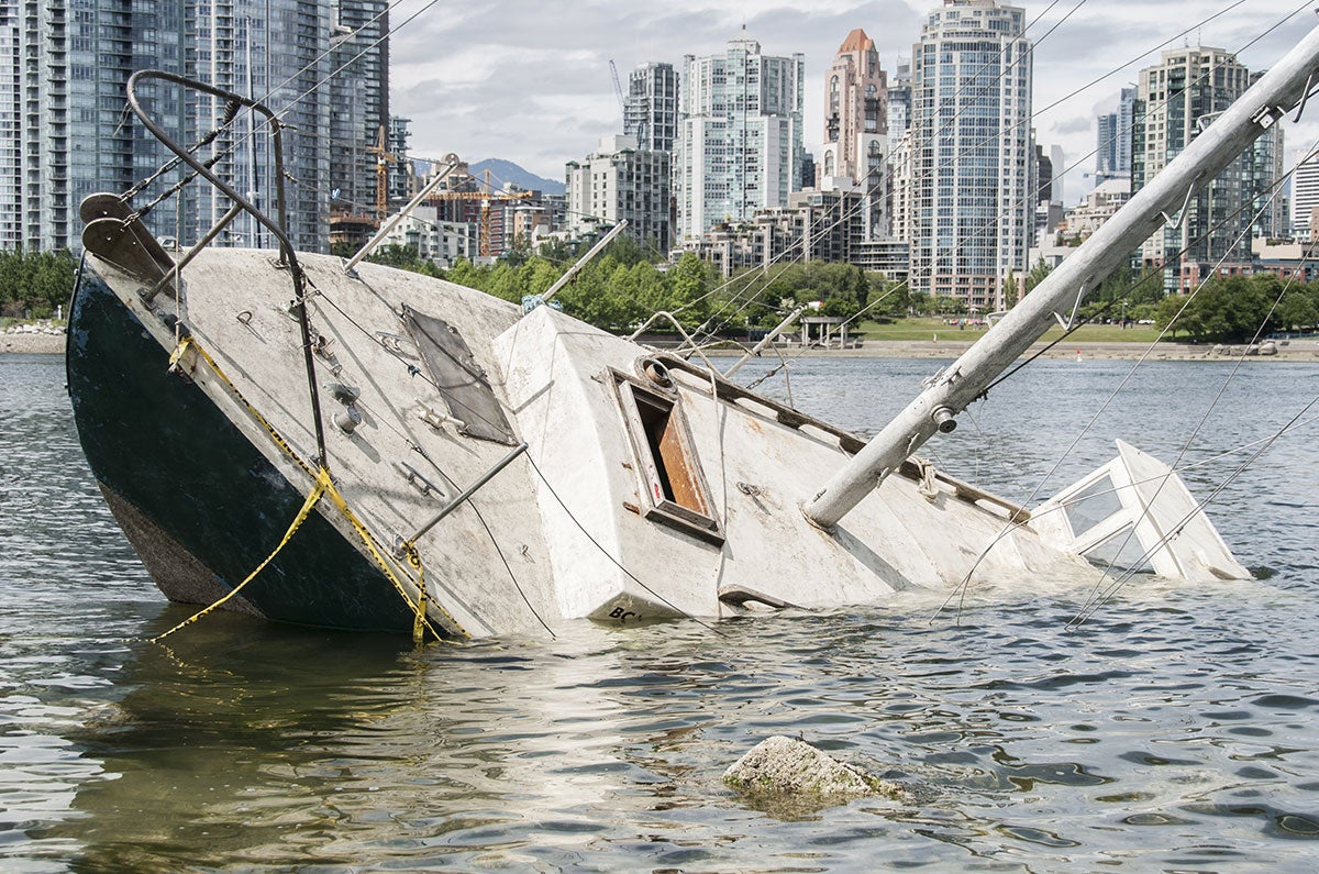 A partially sunken sailboat lists heavily in the water, with a city skyline visible in the background.