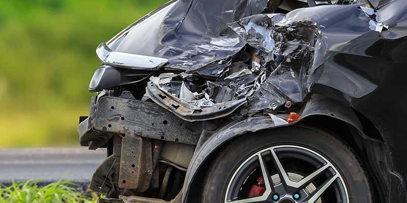 Front left side of a black car with severe crash damage, crumpled hood, bumper, and exposed internal components.