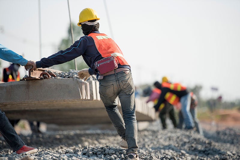 Construction workers in hard hats and safety vests install heavy concrete railroad ties on a rocky track bed.