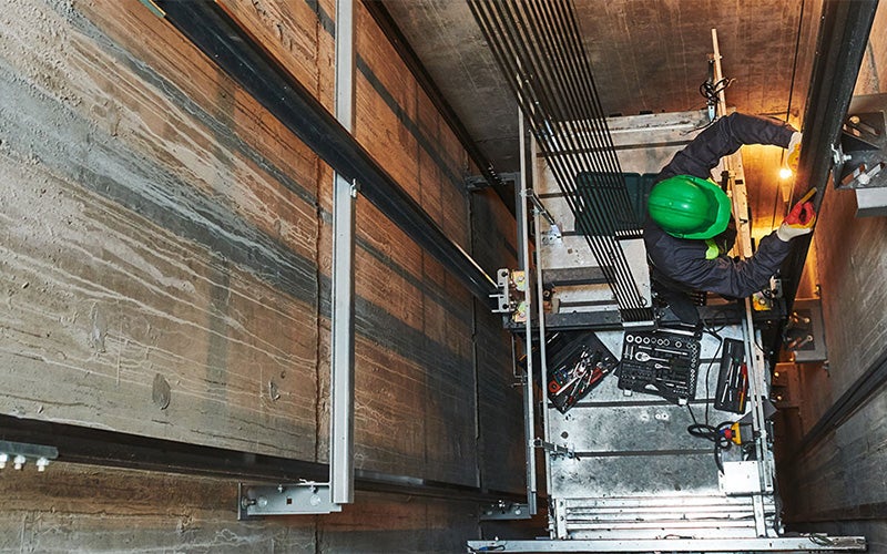 Elevator technician in a green hard hat works on top of an elevator car, seen from above in the shaft.