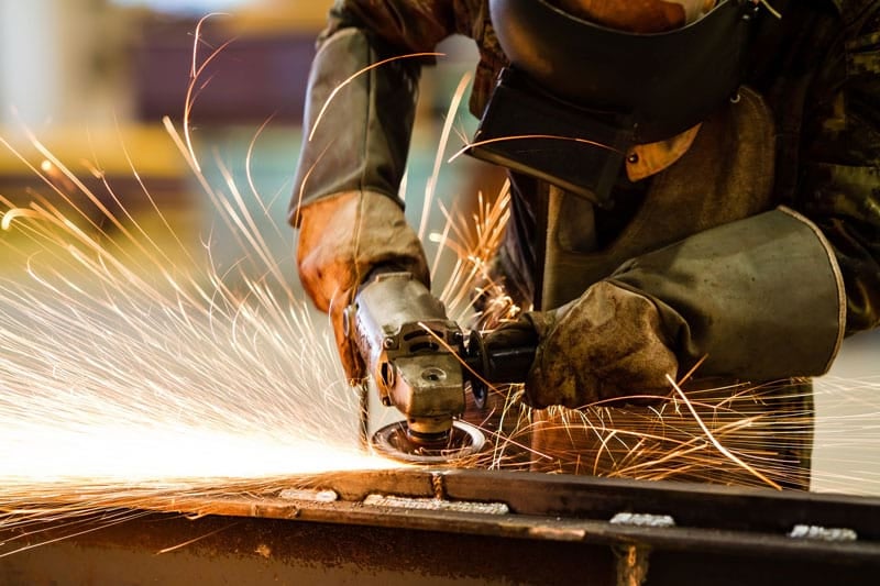 Worker in protective gear using an angle grinder on metal, sending a shower of bright orange sparks across the scene.