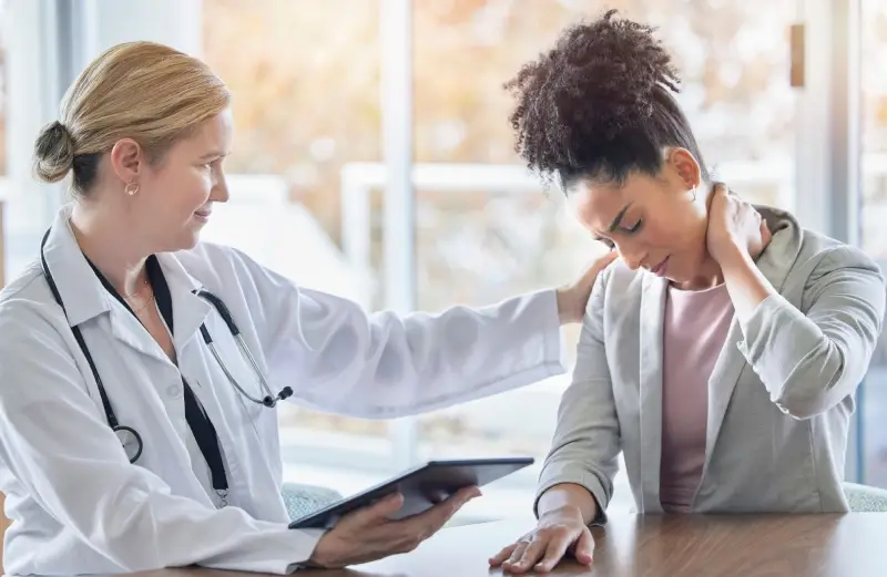 A doctor comforts a distressed patient holding her neck in pain, possibly discussing a diagnosis from a tablet.