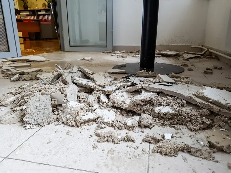 Broken plasterboard and debris cover a tiled, wet floor in a damaged room with an open doorway.