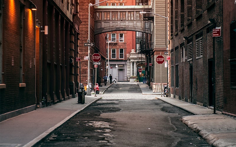 A narrow, dark city alley with tall brick buildings, a skybridge overhead, and multiple stop signs.