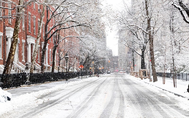 Snow-covered New York City street lined with brownstone buildings and trees during a winter snowfall