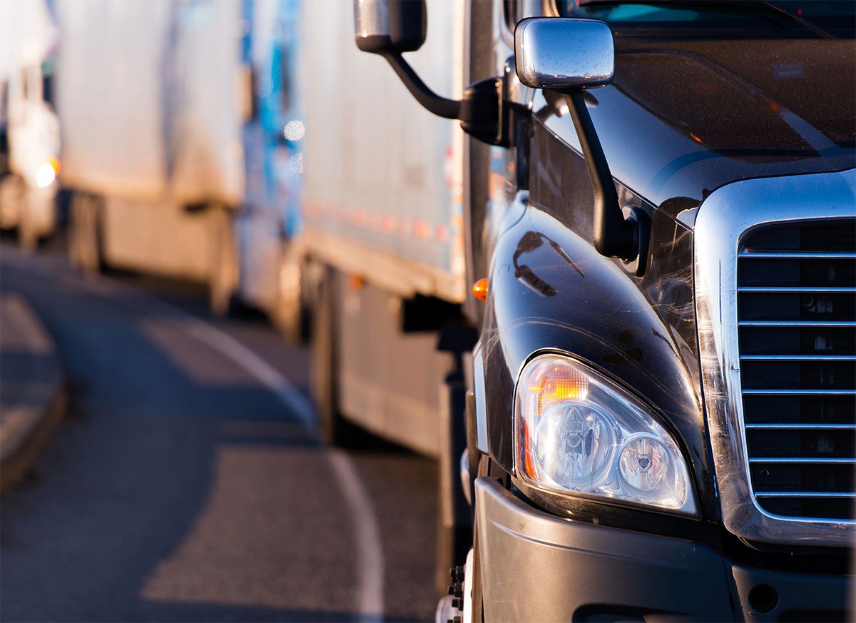 Black semi-truck with a line of other trucks parked or moving slowly on a road at sunset.